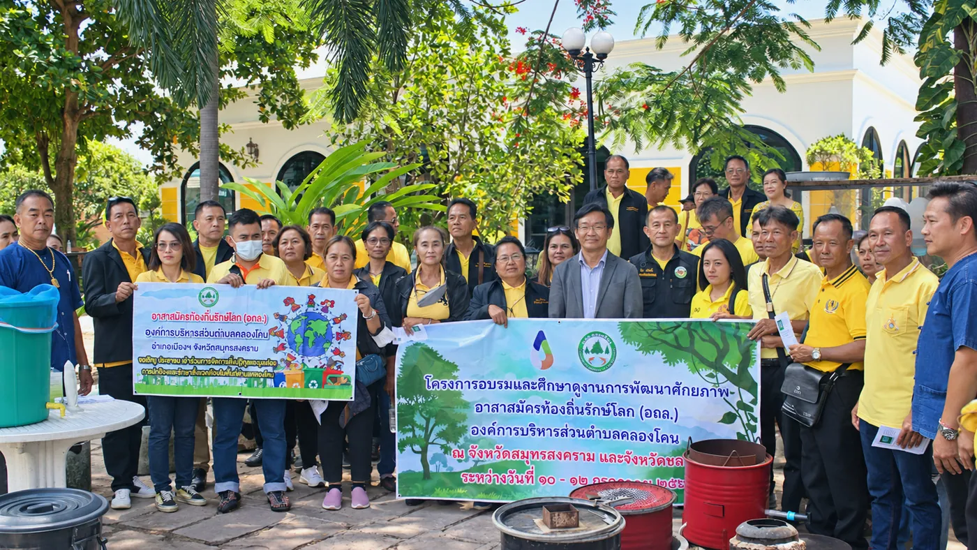 ภาพหมู่ผู้เข้าร่วมโครงการอบรมและศึกษาดูงานการพัฒนาศักยภาพอาสาสมัครท้องถิ่นรักษ์โลก (อถล.) องค์การบริหารส่วนตำบลคลองโคน อำเภอเมืองฯ จังหวัดสมุทรสงคราม ถือป้ายไวนิลโครงการ ด้านหน้ามีถังหมัก ถังน้ำ และเตาชีวมวลตั้งแสดง